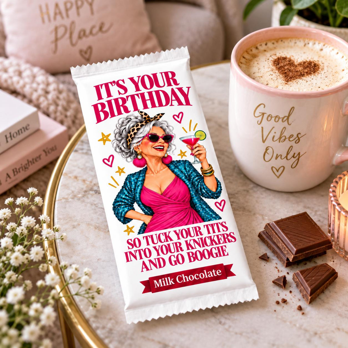A milk chocolate bar wrapped in a light beige  paper sleeve with a colorful retro illustration of a woman and the text 'IT'S YOUR BIRTHDAY  GO BOOGIE', styled on a marble surface next to a mug of coffee and chocolate pieces.