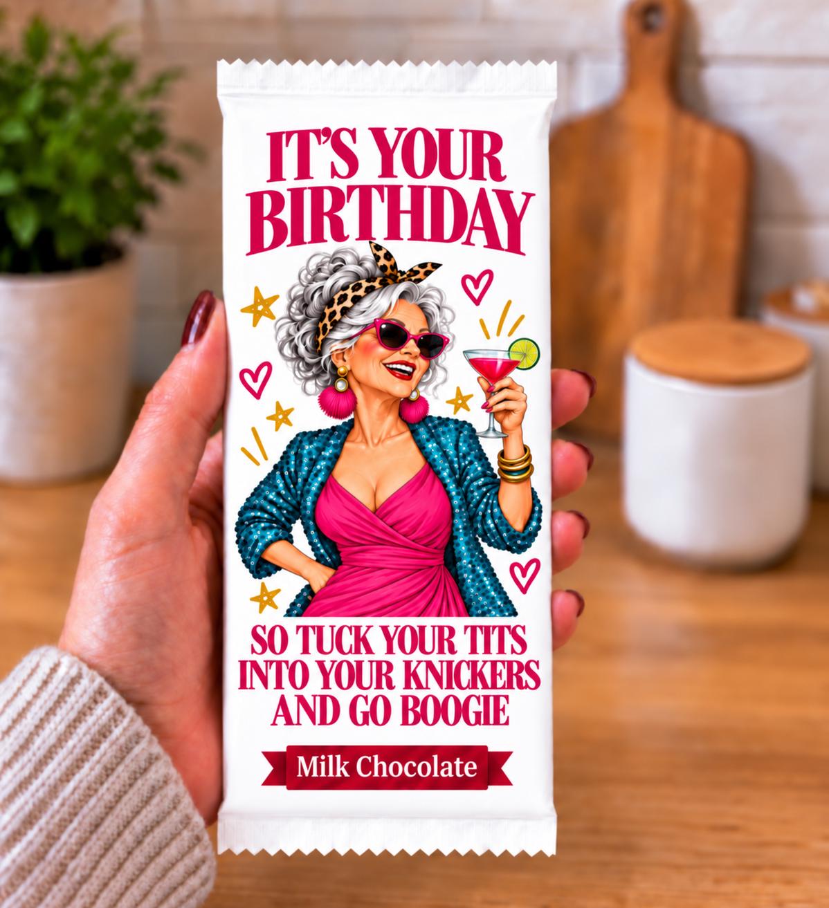 A hand holds a milk chocolate bar wrapper featuring a colorful illustration of a woman and the text 'IT'S YOUR BIRTHDAY  GO BOOGIE' against a blurred kitchen background.