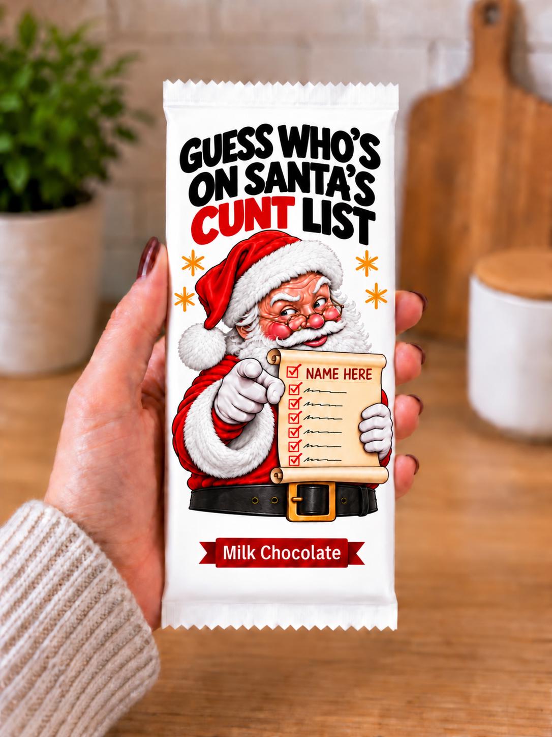 A hand holding a custom printed milk chocolate bar wrapper including chocolate bar featuring a cartoon Santa pointing to a checklist with the text 'GUESS WHO'S ON SANTA'S NAUGHTY LIST' and 'Milk Chocolate' at the bottom, set against a blurred kitchen background.