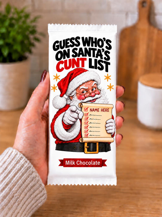A hand holding a custom printed milk chocolate bar wrapper including chocolate bar featuring a cartoon Santa pointing to a checklist with the text 'GUESS WHO'S ON SANTA'S NAUGHTY LIST' and 'Milk Chocolate' at the bottom, set against a blurred kitchen background.