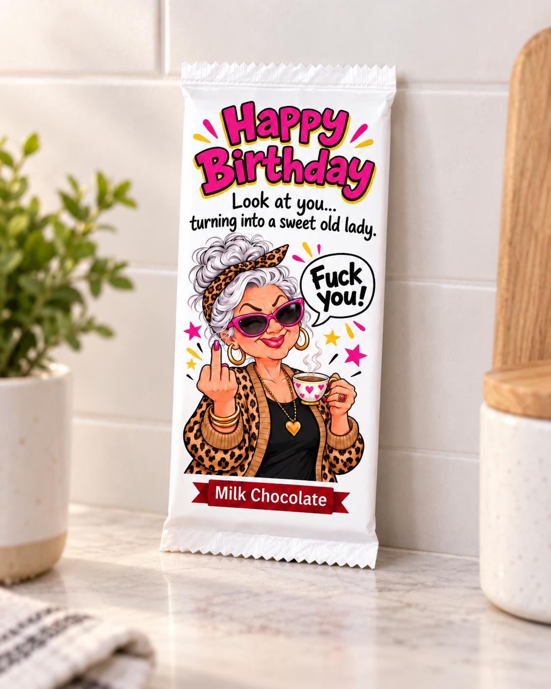 A novelty milk chocolate bar in a white wrapper featuring a cartoon illustration of an angry senior woman and the text 'Happy Birthday' and 'Milk Chocolate', photographed on a kitchen countertop.
